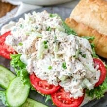 A classic chicken salad on a roll with tomatoes and lettuce next to cucumbers.